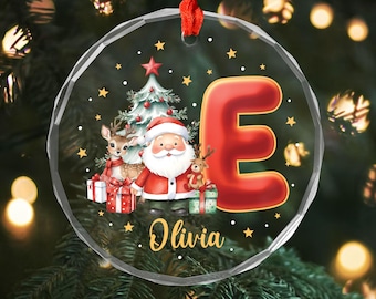 May include: A clear, round Christmas ornament with a red ribbon. The ornament features a festive design with Santa Claus, a reindeer, a Christmas tree, and the letter "E". The name "Olivia" is written in gold.