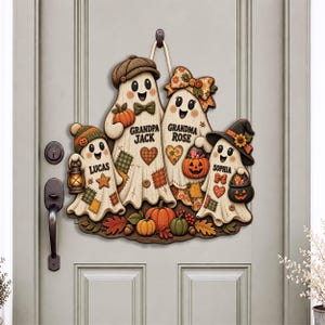 May include: A decorative wooden door hanger featuring four ghost figures labelled Grandpa Jack, Grandma Rose, Lucas, and Sophia. The ghosts are adorned with autumn-themed accessories like hats, bows, pumpkins, and lanterns, set against a light grey door.