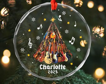 May include: A clear, round Christmas ornament with a red ribbon. The ornament features a Christmas tree made of guitars, musical notes, snowflakes, and stars. The name "Charlotte" and the year "2025" are printed on the ornament.