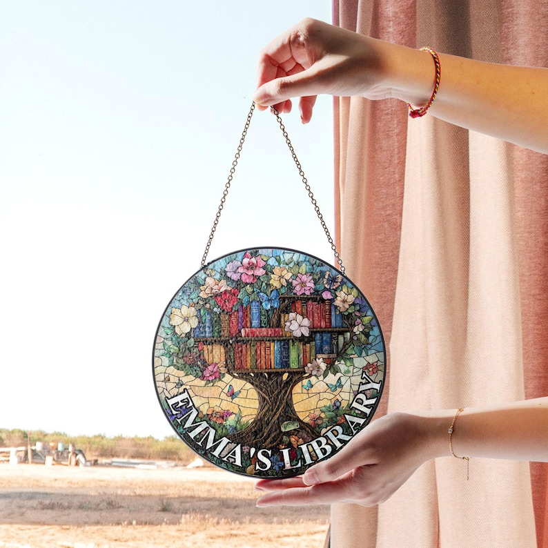 May include: A round stained glass window hanging with a chain. The window features a colorful tree with books on the branches and the text "Emma's Library".