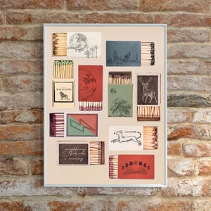 May include: A framed artwork featuring various matchboxes with unique designs. The matchboxes showcase illustrations of a deer, a cityscape, a vinyl record, and a dog. Some boxes have text, including "Marauders" and "anything for a moody". The artwork is displayed against a brick wall.
