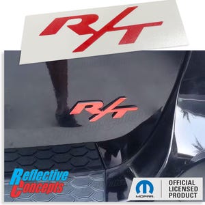 May include: A red and black "R/T" emblem with a reflective finish. The emblem is attached to a black car. The emblem is an official licensed product from Mopar.
