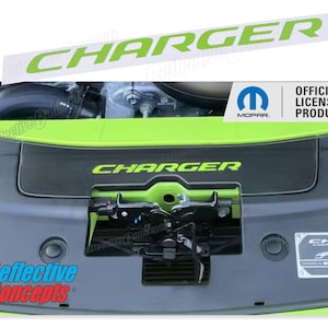 May include: A green and black car part with the word "CHARGER" printed in green on a black background. The part is made of plastic and has a latch mechanism. The image also includes a white sticker with the word "CHARGER" printed in green on a white background. The sticker is an official licensed product.