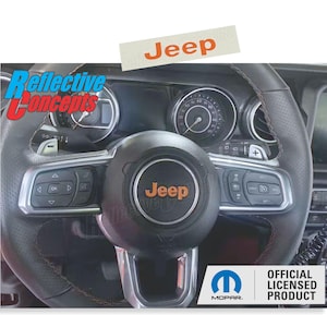 May include: A black leather steering wheel with orange Jeep logo and silver accents. The steering wheel has controls for volume, phone, and cruise control. The steering wheel is labeled with the Jeep and Mopar logos. The text "OFFICIAL LICENSED PRODUCT" is also visible.