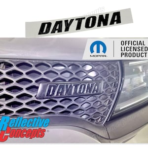 May include: A black and silver car grille with a black "DAYTONA" emblem. The grille is made of a diamond-shaped mesh pattern. The emblem is surrounded by a silver trim. The car grille is part of a vehicle with a silver paint job. The image also includes a white sticker with the word "DAYTONA" in black text and a blue Mopar logo with the text "OFFICIAL LICENSED PRODUCT".