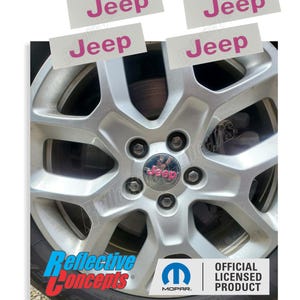 May include: A close-up of a silver car wheel with a black tire. The wheel has a pink Jeep logo in the center and the word "Jeep" in pink on four white labels. The words "Reflective Concepts" and "Mopar" are also visible.