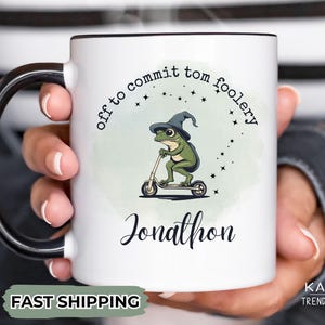 May include: White ceramic mug with a black handle. The mug has a green frog wearing a witch's hat riding a scooter. The text on the mug reads "Off to commit tom foolery Jonathon".