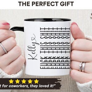 May include: White ceramic mug with a black rim and handle. The mug has a black and white graphic of different sewing stitches and the name "Kellye" written in cursive. The mug is being held by a person's hands.