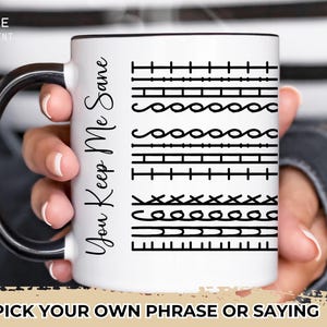 May include: White ceramic mug with a black rim and handle. The mug has black text that says "You Keep Me Sane" and a pattern of different stitches.