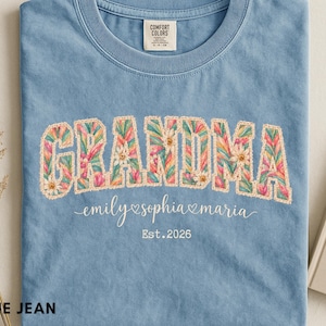 以下が含まれることがあります： ブルーデニムカラーのTシャツで、花柄のGRANDMAという文字と白いステッチの輪郭が特徴です。文字の下には、白い筆記体でemily、sophia、mariaという名前とハートのアイコンが描かれ、その下にEst. 2026というテキストがあります。