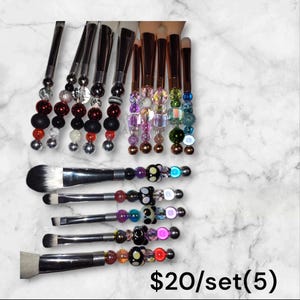 K&ouml;nnte beinhalten: Set aus f&uuml;nf Make-up-Pinseln mit bunten Perlengriffen. Die Pinsel haben silberne Metallgriffe und schwarze, wei&szlig;e und regenbogenfarbene Perlen. Der Text "$20/set(5)" ist im Bild sichtbar.