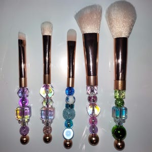 K&ouml;nnte beinhalten: Satz mit sechs Make-up-Pinseln mit ros&eacute;goldenen Griffen und bunten Perlenakzenten. Die Pinsel haben verschiedene Formen und Gr&ouml;&szlig;en f&uuml;r verschiedene Make-up-Anwendungen.
