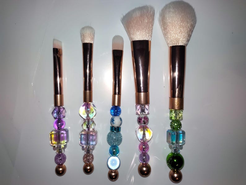 K&ouml;nnte beinhalten: Satz mit sechs Make-up-Pinseln mit ros&eacute;goldenen Griffen und bunten Perlenakzenten. Die Pinsel haben verschiedene Formen und Gr&ouml;&szlig;en f&uuml;r verschiedene Make-up-Anwendungen.