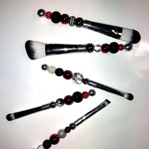 K&ouml;nnte beinhalten: Satz mit f&uuml;nf Make-up-Pinseln mit schwarzen und roten Perlengriffen. Die Pinsel haben silberne Metallfassungen und wei&szlig;e Borsten.