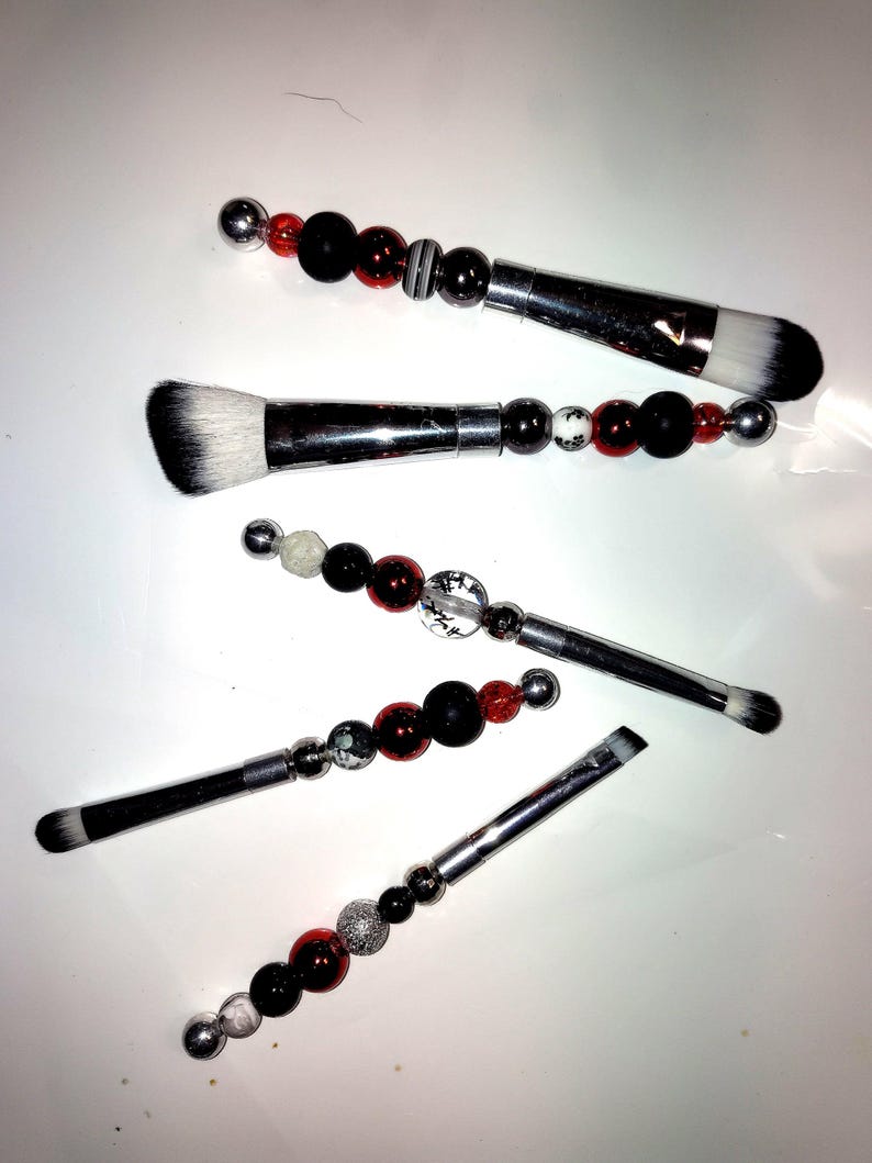 K&ouml;nnte beinhalten: Satz mit f&uuml;nf Make-up-Pinseln mit schwarzen und roten Perlengriffen. Die Pinsel haben silberne Metallfassungen und wei&szlig;e Borsten.