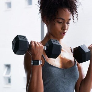 Op de afbeelding: Een vrouw in een grijs tanktop en een witte smartwatch tilt twee zwarte dumbbells tijdens een training.