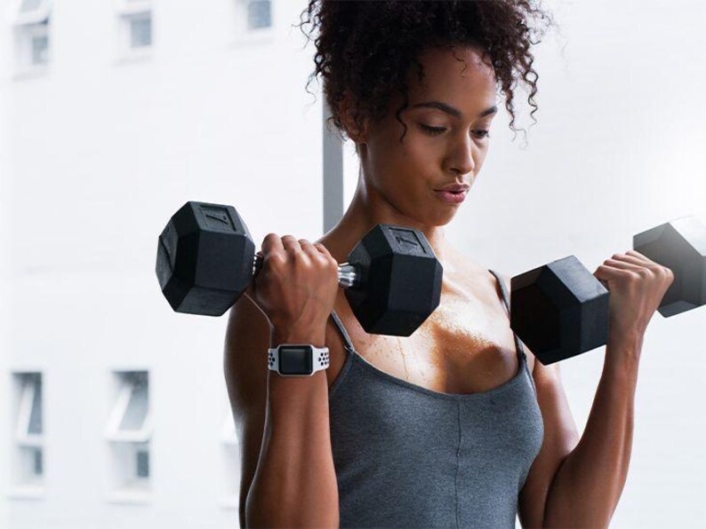 Op de afbeelding: Een vrouw in een grijs tanktop en een witte smartwatch tilt twee zwarte dumbbells tijdens een training.