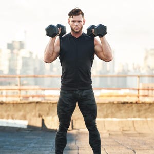May include: A man wearing a black sleeveless shirt and camouflage pants is lifting two black dumbbells.