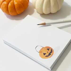 May include: A white notepad with a small orange pumpkin jack-o'-lantern illustration. The notepad is on a white surface with a pencil and two pumpkins.