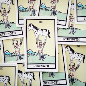 Puede incluir: Un conjunto de cartas del tarot con la carta de la Fuerza. La carta muestra a una mujer levantando un caballo manchado. La palabra "STRENGTH" está en la parte inferior de la carta. El fondo es de color amarillo claro.