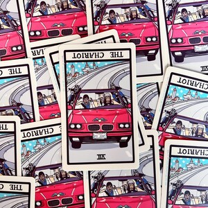 May include: A set of tarot cards featuring the Chariot card. The card depicts a pink car with three people inside driving on a winding road. The card number VII is in the bottom right corner. The card is surrounded by other tarot cards.