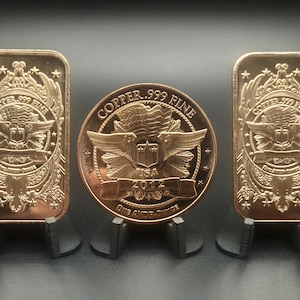 May include: Two rectangular copper bars and one circular copper coin, each stamped with COPPER .999 FINE, an eagle emblem, and the text ONE AVOIR OUNCE. The central coin also features the year 2012 and USA. All items are displayed on black stands.