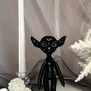 May include: A black felt doll with gold embroidery, featuring a crescent moon and star details on the head and body. The doll has large ears and a slender frame, with delicate stitching. The doll is a unique handmade item.