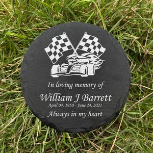 May include: A round, black slate memorial stone with white engraved racing flags and a race car design. The text reads "In loving memory of William J Barrett, April 04, 1950 - June 24, 2021, Always in my heart."