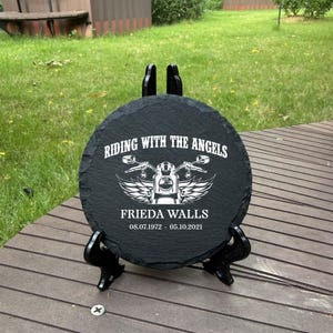 May include: A circular black slate memorial plaque featuring a white engraved graphic of a motorcycle with wings. The text reads RIDING WITH THE ANGELS, the name FRIEDA WALLS, and the dates 08.07.1972 - 05.10.2021. It rests on a black stand.