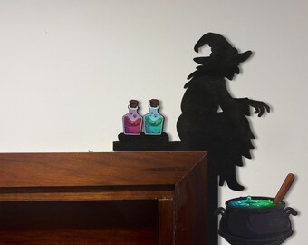 May include: A black silhouette of a witch wearing a pointed hat, sitting on a ledge next to two small glass bottles containing pink and blue liquids, with a black cauldron filled with bubbling green liquid and a wooden stirring spoon nearby.