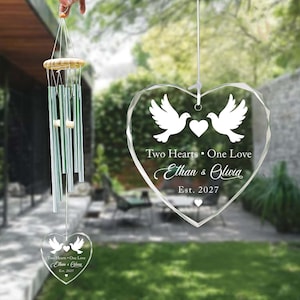 May include: A clear, heart-shaped crystal ornament featuring two white doves and the inscription "Two Hearts • One Love Ethan & Olivia Est. 2027." A wind chime with silver tubes and a wooden top is also visible.