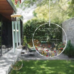 Pode incluir: Um sino de vento memorial com tubos transparentes e um topo de madeira. Um ornamento circular transparente com detalhes florais e o texto "A Father's Love Is Forever Rooted In Our Hearts" e um nome e datas também é visível.