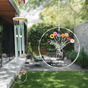 May include: A clear acrylic wind chime with a wooden top and silver-colored chimes. A round acrylic ornament features a floral bouquet design with the text "Granny We Love You". The wind chime is hanging outdoors.