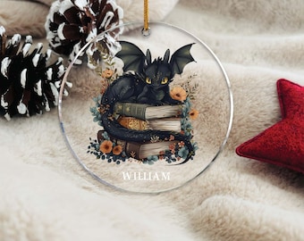May include: A clear, round ornament featuring a black dragon resting on a stack of books. The dragon has yellow eyes and is surrounded by flowers. The name "WILLIAM" is printed at the bottom. A gold string is attached for hanging.