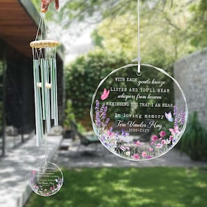 May include: A memorial wind chime with a clear, circular acrylic pendant featuring floral and butterfly designs. The text reads "WITH EACH gentle breeze LISTEN and YOU'LL HEAR whispers from heaven... In loving memory Tere Vander May 1990-2025."