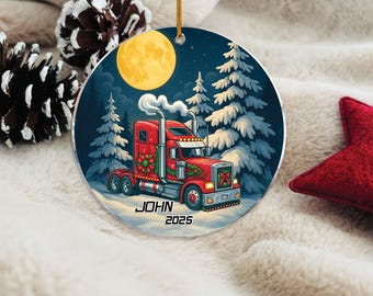May include: A round Christmas ornament featuring a red semi-truck with a winter scene. The truck has holiday decorations and the text "JOHN 2025". A full moon and snow-covered trees are in the background. The ornament is hanging by a gold string.