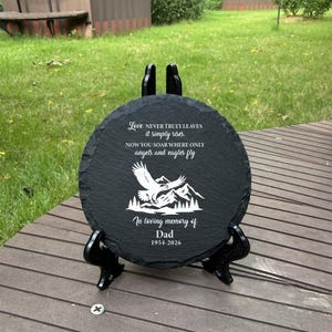 May include: A circular black slate memorial plaque featuring a white engraved eagle soaring over mountain peaks. The text reads: Love NEVER TRULY LEAVES it simply rises. NOW YOU SOAR WHERE ONLY angels and eagles fly. In loving memory of Dad 1954-2026.