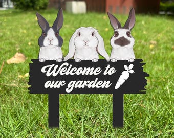 May include: A black metal garden stake featuring three rabbit illustrations above the text "Welcome to our garden" and a carrot graphic. The rabbits are white with black and brown markings. The stake is set in green grass.