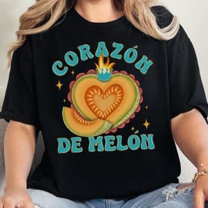 Op de afbeelding: Zwart T-shirt met een afbeelding van een hartvormige cantaloupemeloen met een kroon en de tekst "CORAZ&Oacute;N DE MEL&Oacute;N" in blauw.