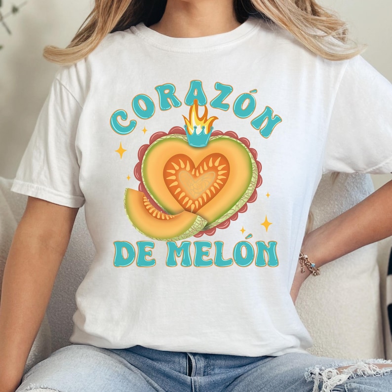 Op de afbeelding: Wit T-shirt met een afbeelding van een hartvormige cantaloupe met een kroon erop. De tekst "CORAZ&Oacute;N DE MEL&Oacute;N" is in blauwe letters geschreven.