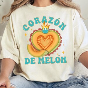 Op de afbeelding: Een wit T-shirt met een afbeelding van een hartvormige cantaloupemeloen met een kroon en vlammen. De tekst "CORAZ&Oacute;N DE MEL&Oacute;N" staat in blauw op het shirt.