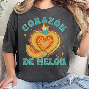 Op de afbeelding: Een donkergrijs T-shirt met een vintage-stijl grafische afbeelding van een hartvormige cantaloupe met een kroon en de tekst "CORAZ&Oacute;N DE MEL&Oacute;N".