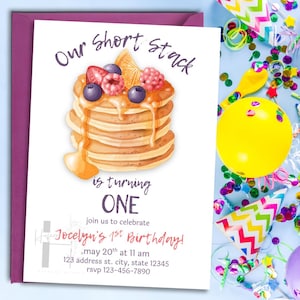 Puede incluir: Una invitación de cumpleaños con una ilustración de panqueques con bayas y jarabe. El texto dice "Our Short Stack is turning ONE" e incluye detalles de la fiesta. La invitación está en una tarjeta blanca con un sobre morado.