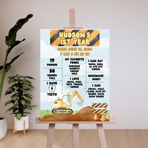 以下が含まれることがあります： 青い空を背景に、黄色の建設車両。 「Hudson's 1st Year」というテキストと、子供の最初の年の詳細、体重、身長、好きな食べ物、マイルストーンなどが書かれたチャート。