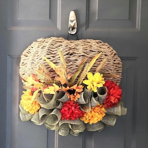 May include: A fall-themed door wreath with a burlap base and a variety of yellow, orange, and red flowers. The wreath is decorated with burlap ribbon and wheat stalks.