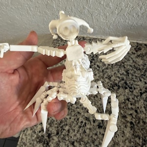 Puede incluir: Una figura blanca, articulada, impresa en 3D, con forma de escorpi&oacute;n. La figura tiene una cabeza con un sombrero de buf&oacute;n, un cuerpo segmentado y m&uacute;ltiples extremidades articuladas con puntas afiladas. La figura se sostiene en una mano.