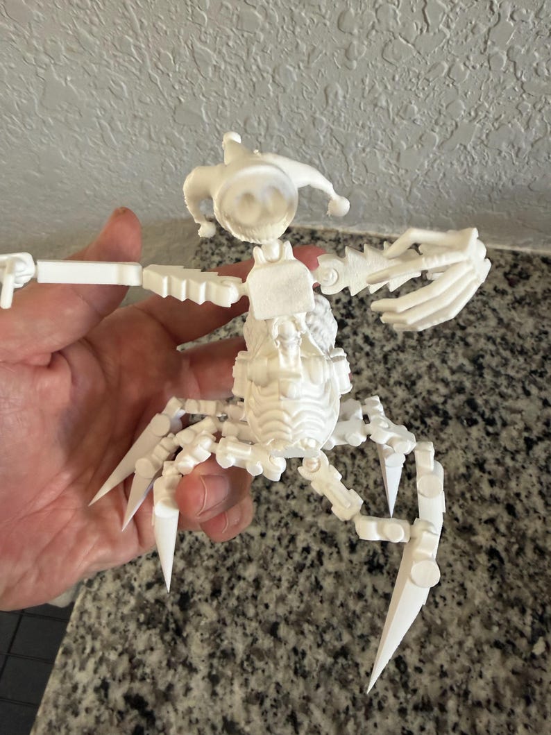 Puede incluir: Una figura blanca, articulada, impresa en 3D, con forma de escorpi&oacute;n. La figura tiene una cabeza con un sombrero de buf&oacute;n, un cuerpo segmentado y m&uacute;ltiples extremidades articuladas con puntas afiladas. La figura se sostiene en una mano.