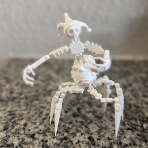 Puede incluir: Una escultura blanca impresa en 3D de una figura esquel&eacute;tica parecida a un buf&oacute;n. Tiene un gorro de buf&oacute;n, un cuerpo segmentado y m&uacute;ltiples patas de ara&ntilde;a. La figura est&aacute; posando con un brazo extendido.
