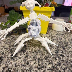 Könnte beinhalten: Eine weiße, bewegliche Skelettfigur mit schädelartigem Kopf, einem durchsichtigen Brustteil und beweglichen Gliedmaßen. Die Figur steht auf einer gesprenkelten Arbeitsplatte, im Hintergrund ein gelber Blumentopf.