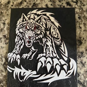 May include: A black rectangular plaque featuring a white, stylized wolf design. The wolf is depicted in a snarling pose with red eyes and intricate tribal patterns. The artwork is a detailed, three-dimensional relief.